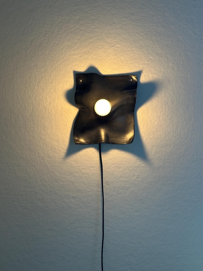 May include: A wall-mounted lamp featuring a dark, wavy, square-shaped backplate with a central circular light bulb. The fixture emits a warm glow, casting a distinct shadow against the wall, and includes a thin black power cord extending downward.