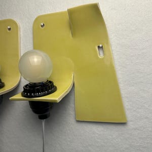 May include: A wall-mounted light fixture featuring a pale yellow ceramic backplate with an irregular, folded shape. A black socket holds a round, frosted white light bulb, and a thin white electrical cord extends downward from the base.