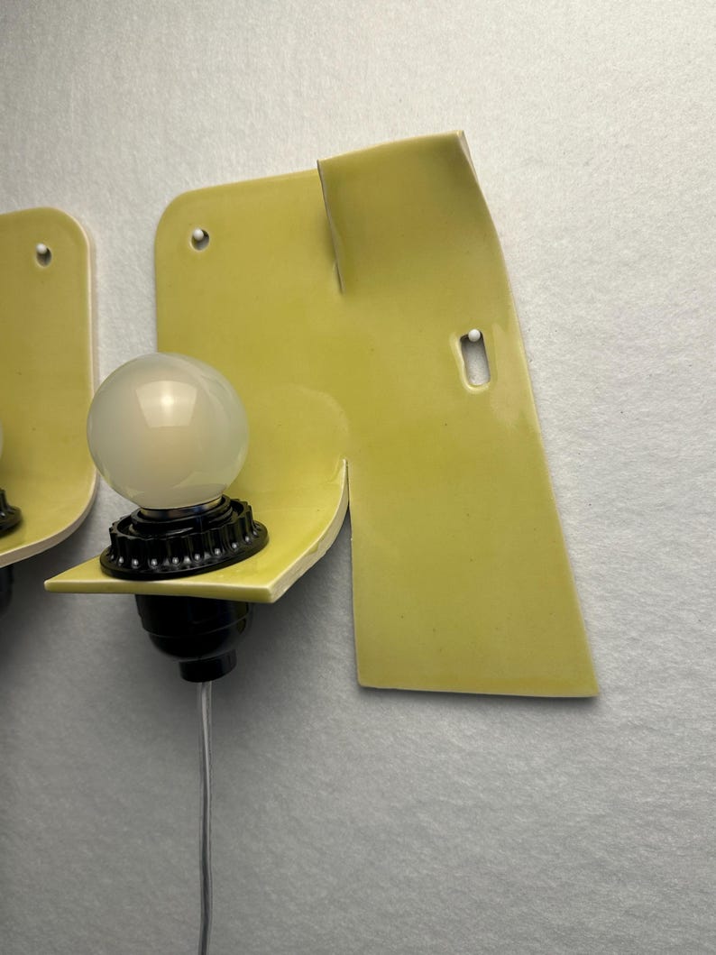 May include: A wall-mounted light fixture featuring a pale yellow ceramic backplate with an irregular, folded shape. A black socket holds a round, frosted white light bulb, and a thin white electrical cord extends downward from the base.