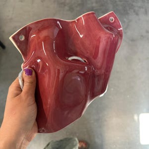 Puede incluir: Una maceta de pared de cer&aacute;mica en color rojo intenso brillante con una forma org&aacute;nica ondulada. La pieza cuenta con dos orificios circulares en las esquinas superiores para su montaje y una abertura central, con un acabado liso y reflectante.