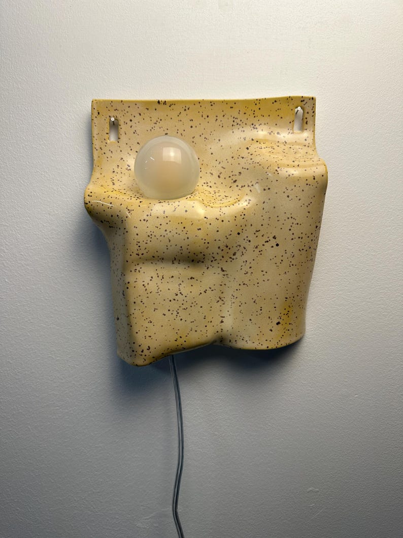 May include: A wall-mounted ceramic light fixture with a speckled cream and yellow glaze. The sculptural form features an irregular, organic shape with a recessed circular opening holding a single frosted glass globe bulb and two top mounting holes.