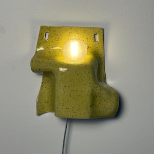 Puede incluir: Un aplique de pared de cer&aacute;mica con esmalte amarillo p&aacute;lido y motas negras. La l&aacute;mpara presenta una forma fluida y drapeada con dos orificios de montaje rectangulares en la parte superior y una bombilla esf&eacute;rica brillante en el centro.