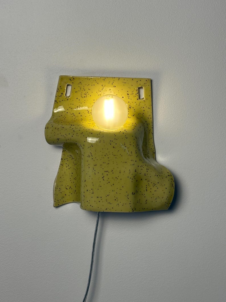 Puede incluir: Un aplique de pared de cer&aacute;mica con esmalte amarillo p&aacute;lido y motas negras. La l&aacute;mpara presenta una forma fluida y drapeada con dos orificios de montaje rectangulares en la parte superior y una bombilla esf&eacute;rica brillante en el centro.