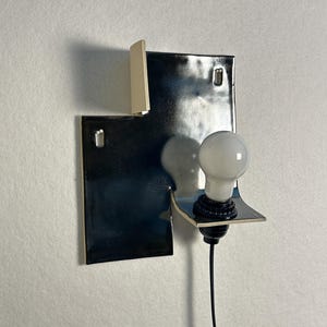 Pode incluir: Uma lumin&aacute;ria de parede moderna com um design geom&eacute;trico preto brilhante. Possui uma l&acirc;mpada branca e um cabo preto. A lumin&aacute;ria tem uma forma art&iacute;stica &uacute;nica e &eacute; montada em uma parede branca.