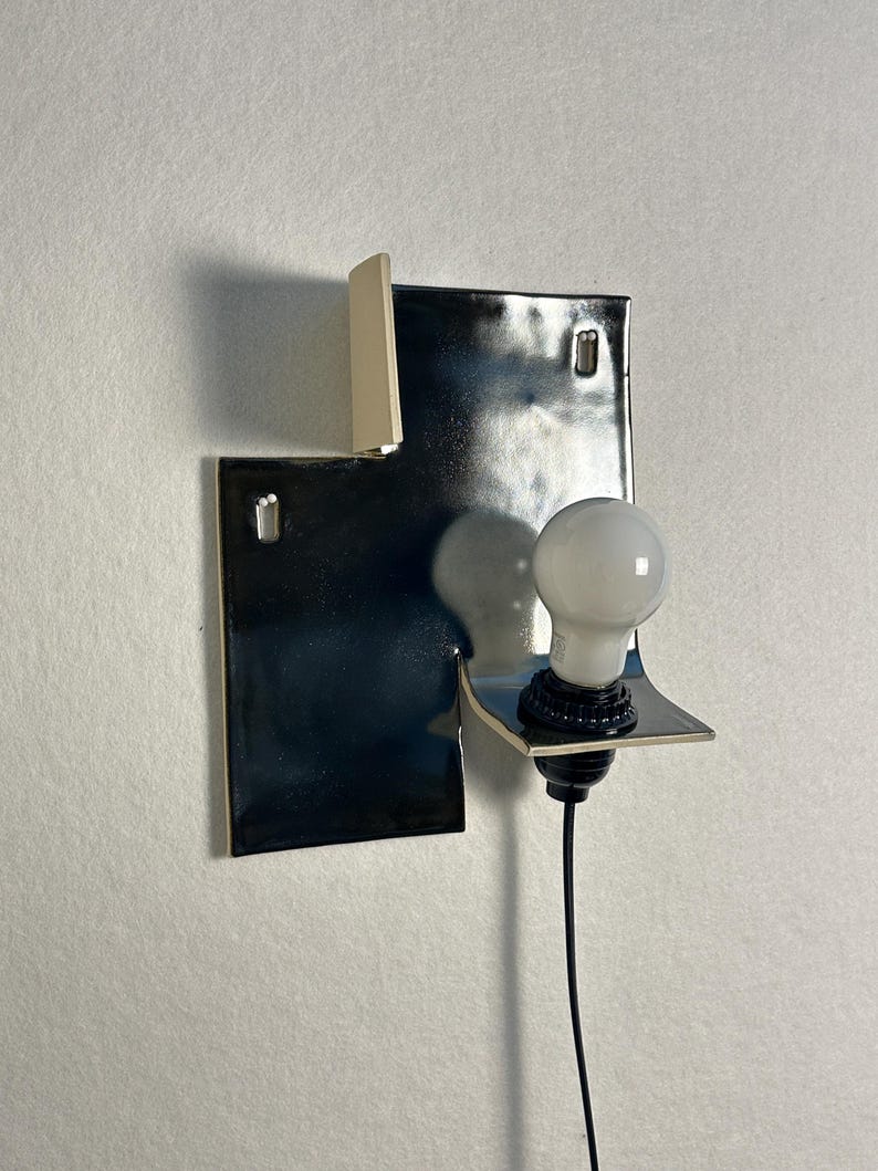 Pode incluir: Uma lumin&aacute;ria de parede moderna com um design geom&eacute;trico preto brilhante. Possui uma l&acirc;mpada branca e um cabo preto. A lumin&aacute;ria tem uma forma art&iacute;stica &uacute;nica e &eacute; montada em uma parede branca.