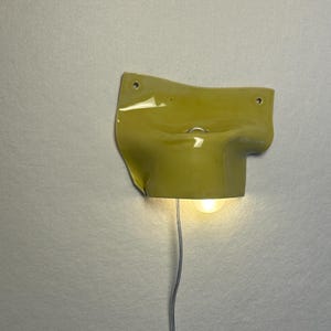 May include: A yellow ceramic wall sconce with a curved design. The sconce has a small light bulb and a silver wire. The fixture is mounted on a white wall. The sconce has three small holes.