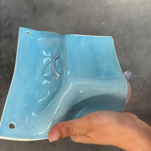 K&ouml;nnte beinhalten: Eine hellblaue Wandleuchte aus Keramik mit gl&auml;nzender Oberfl&auml;che, die eine geschwungene, rechteckige Form aufweist, mit zwei kreisf&ouml;rmigen Befestigungsl&ouml;chern an den oberen und unteren Kanten sowie einem zentralen Metallring.