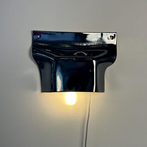 May include: A glossy black ceramic wall sconce with a wide, curved top section featuring two mounting holes and a central opening. A single warm-toned light bulb is positioned underneath the fixture, which is connected to a thin white power cord.