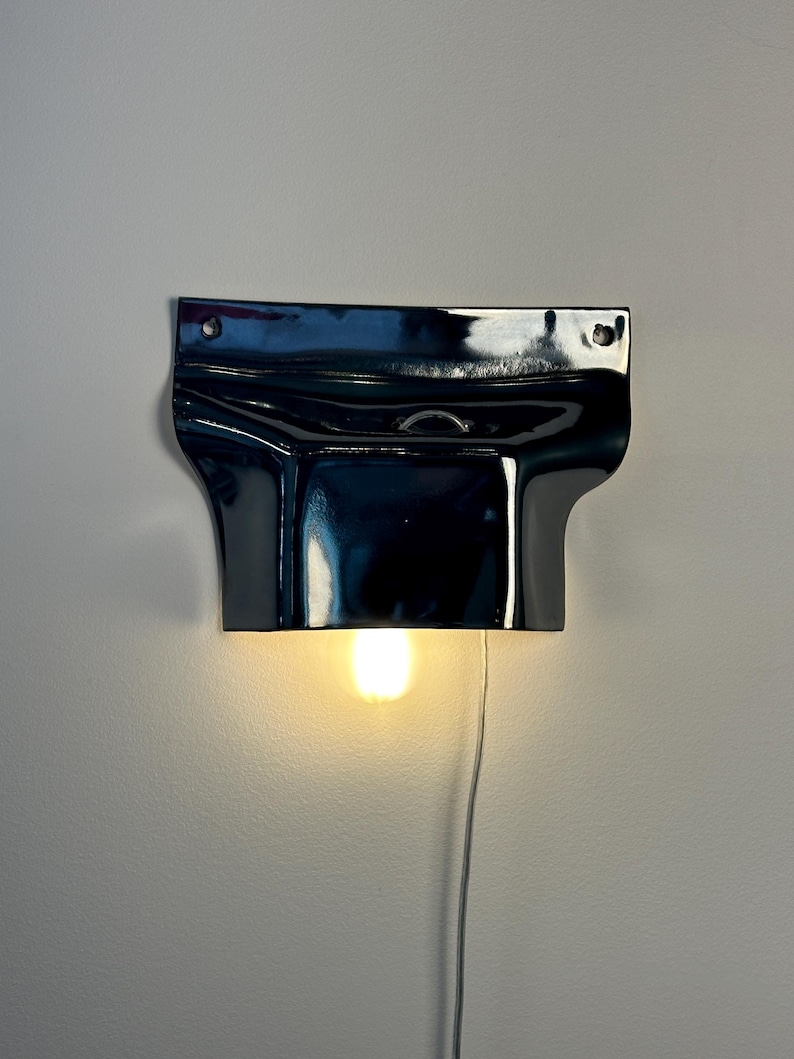 May include: A glossy black ceramic wall sconce with a wide, curved top section featuring two mounting holes and a central opening. A single warm-toned light bulb is positioned underneath the fixture, which is connected to a thin white power cord.