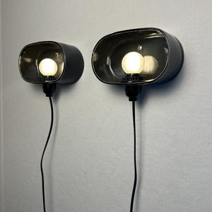 May include: Two black, oval wall sconces, each with a light bulb inside. They are mounted on a light grey wall, with a black cord hanging from each. The bulbs are lit, emitting a warm glow. The sconces are a modern lighting fixture.