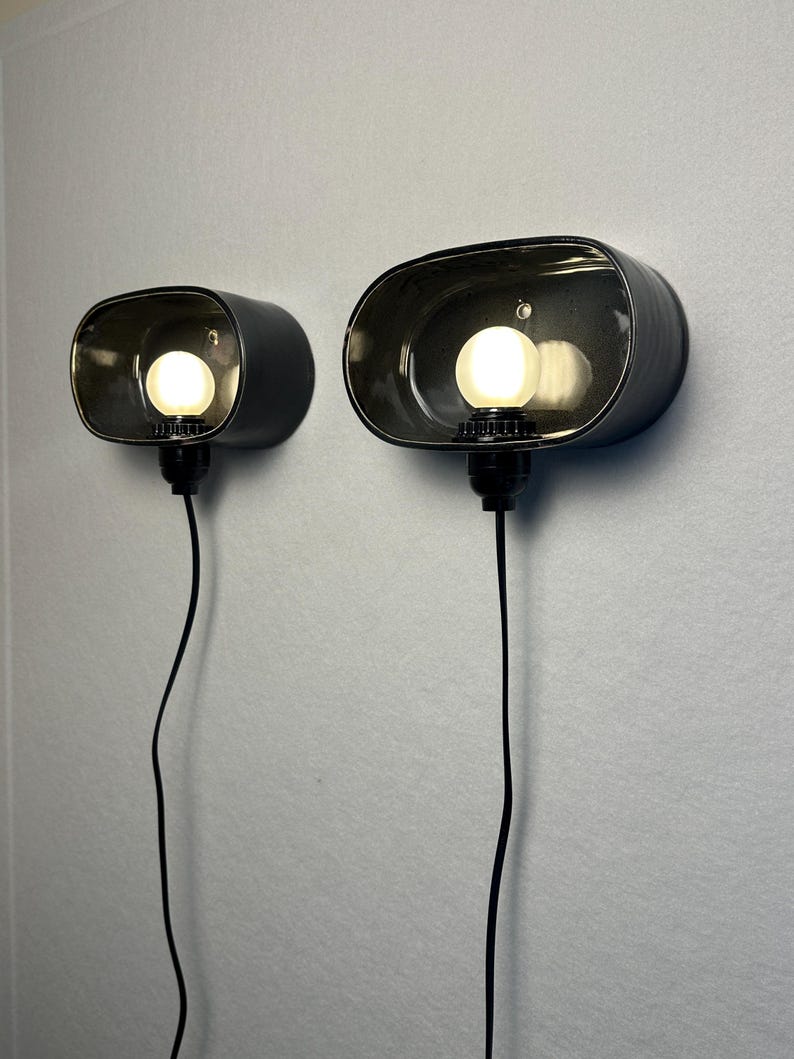 May include: Two black, oval wall sconces, each with a light bulb inside. They are mounted on a light grey wall, with a black cord hanging from each. The bulbs are lit, emitting a warm glow. The sconces are a modern lighting fixture.