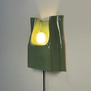 May include: A sage green ceramic wall sconce with a glossy finish and a curved, organic shape. The fixture features a central cutout housing a glowing, warm-toned light bulb. A black power cord extends from the bottom of the wall-mounted light.