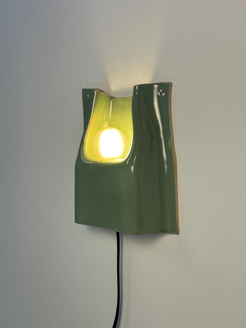 May include: A sage green ceramic wall sconce with a glossy finish and a curved, organic shape. The fixture features a central cutout housing a glowing, warm-toned light bulb. A black power cord extends from the bottom of the wall-mounted light.