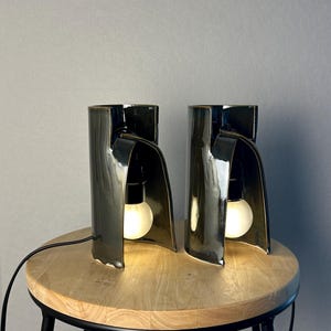 Puede incluir: Dos l&aacute;mparas de mesa de cer&aacute;mica en color verde oliva oscuro con acabado brillante. Cada l&aacute;mpara presenta una estructura cil&iacute;ndrica curva con una secci&oacute;n frontal abierta que alberga una bombilla blanca encendida. Un cable negro sale de la base.
