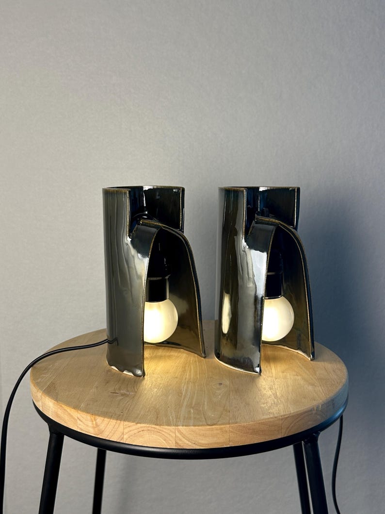 Puede incluir: Dos l&aacute;mparas de mesa de cer&aacute;mica en color verde oliva oscuro con acabado brillante. Cada l&aacute;mpara presenta una estructura cil&iacute;ndrica curva con una secci&oacute;n frontal abierta que alberga una bombilla blanca encendida. Un cable negro sale de la base.