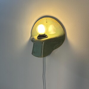 Puede incluir: Un aplique de pared con una placa trasera de cer&aacute;mica semicircular en color verde claro moteado, que cuenta con un estante saliente que sostiene una bombilla redonda brillante. Un cable el&eacute;ctrico blanco cuelga verticalmente de la base.