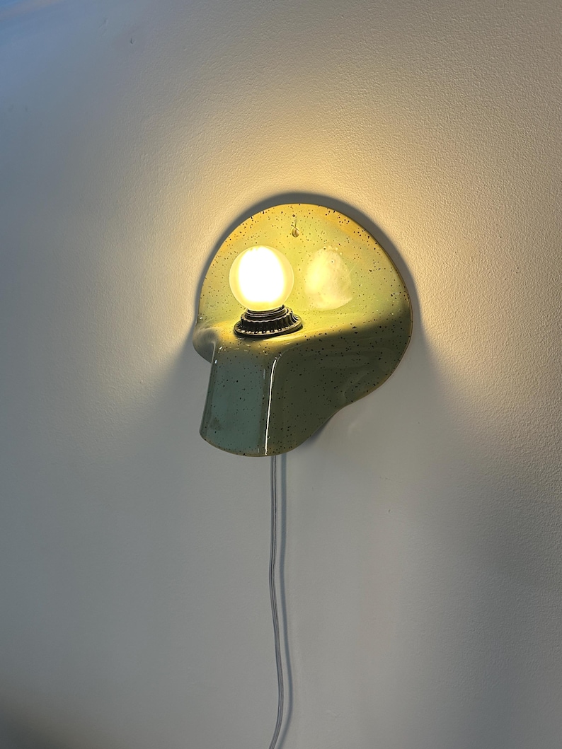 Puede incluir: Un aplique de pared con una placa trasera de cer&aacute;mica semicircular en color verde claro moteado, que cuenta con un estante saliente que sostiene una bombilla redonda brillante. Un cable el&eacute;ctrico blanco cuelga verticalmente de la base.
