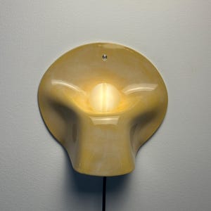 May include: A wall-mounted lamp featuring a glossy, pale yellow ceramic shade with a fluid, organic, and undulating shape. A single, round, glowing white light bulb is positioned in the center of the shade, and a black power cord extends downward.