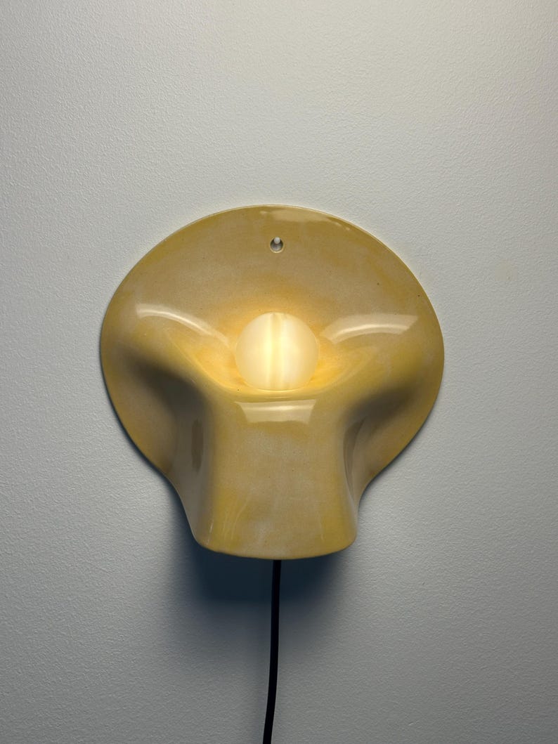May include: A wall-mounted lamp featuring a glossy, pale yellow ceramic shade with a fluid, organic, and undulating shape. A single, round, glowing white light bulb is positioned in the center of the shade, and a black power cord extends downward.
