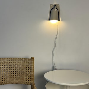 May include: A wall-mounted lamp with a metallic, bell-shaped shade that emits a warm, downward-facing light. The fixture features a thin, transparent power cord that hangs vertically against the wall, connecting to a plug near the floor.