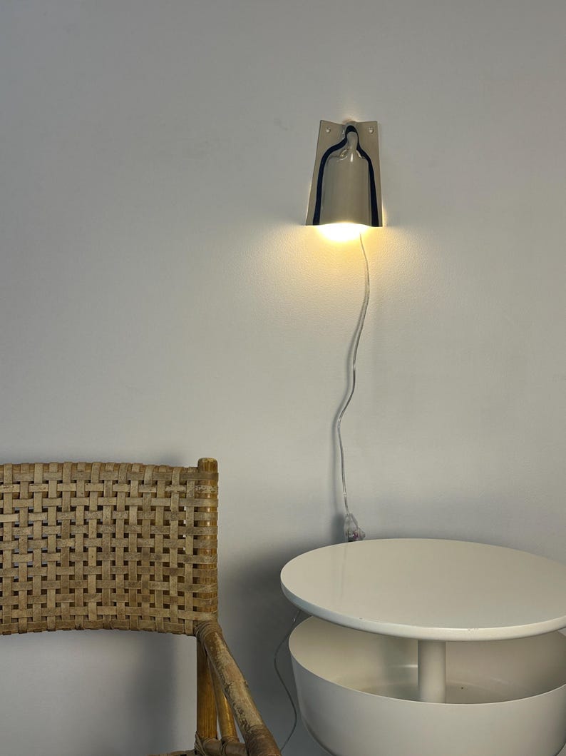 May include: A wall-mounted lamp with a metallic, bell-shaped shade that emits a warm, downward-facing light. The fixture features a thin, transparent power cord that hangs vertically against the wall, connecting to a plug near the floor.