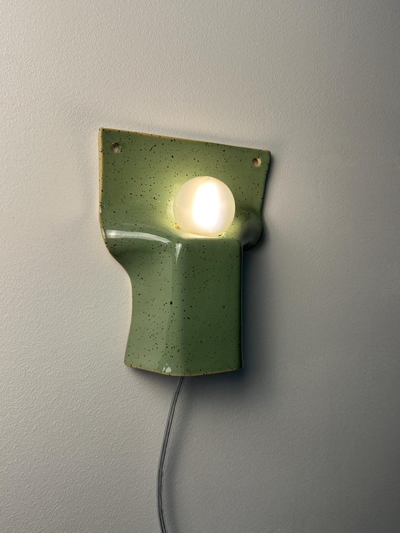 May include: A wall-mounted light fitting featuring a sage green ceramic base with dark speckles and a smooth, glossy finish. The base has a curved, moulded shape with two small holes at the top corners and a single glowing white spherical bulb.