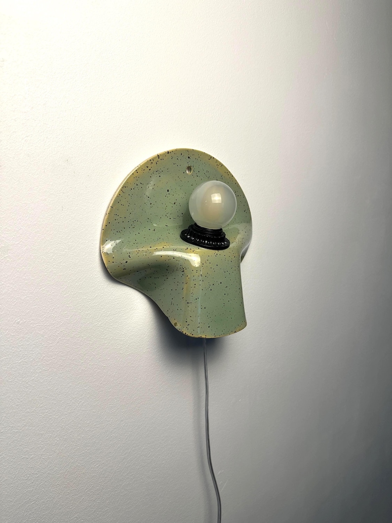 Puede incluir: Un aplique de pared con una base de cer&aacute;mica curva de color verde claro moteado y acabado liso. Una bombilla peque&ntilde;a, redonda y de vidrio esmerilado blanco est&aacute; asegurada en un casquillo negro centrado en la forma cer&aacute;mica, con un cable fino.