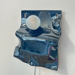 May include: A wall-mounted lamp featuring a reflective, metallic blue, wavy ceramic backplate with an irregular, organic shape. A single round, frosted white light bulb is mounted in a dark socket centered on the upper portion of the surface.