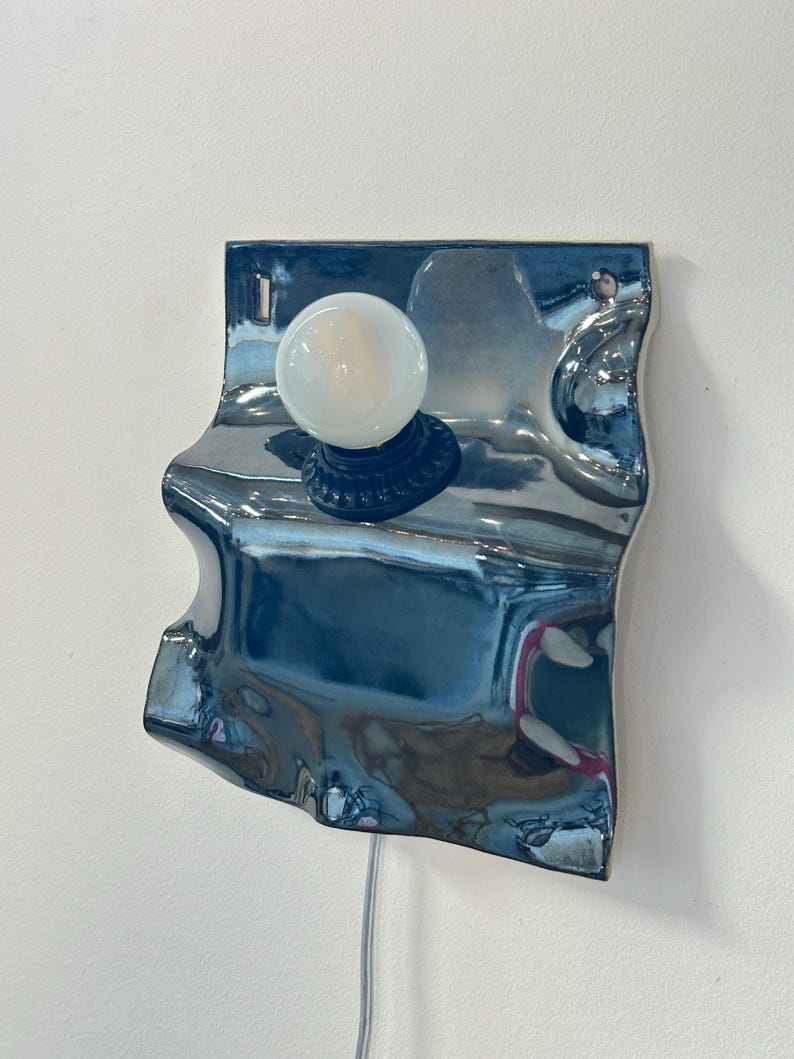 May include: A wall-mounted lamp featuring a reflective, metallic blue, wavy ceramic backplate with an irregular, organic shape. A single round, frosted white light bulb is mounted in a dark socket centered on the upper portion of the surface.