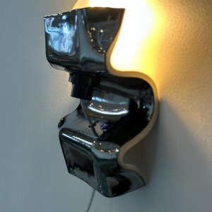 May include: A wall-mounted lamp featuring a glossy, black, S-shaped ceramic body with a reflective surface. The fixture holds a single exposed light bulb that emits a warm, golden glow, casting light upward and downward against the wall surface.