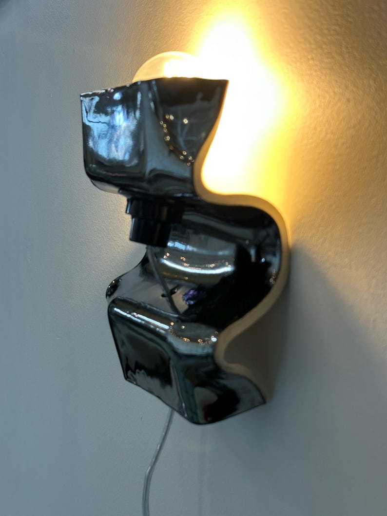 May include: A wall-mounted lamp featuring a glossy, black, S-shaped ceramic body with a reflective surface. The fixture holds a single exposed light bulb that emits a warm, golden glow, casting light upward and downward against the wall surface.