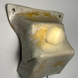 May include: A wall-mounted ceramic light fixture with a glossy, mottled glaze in shades of cream, beige, and yellow. The rectangular, slightly curved form features two mounting holes at the top corners and a single glowing round light bulb.