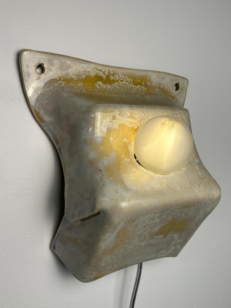 May include: A wall-mounted ceramic light fixture with a glossy, mottled glaze in shades of cream, beige, and yellow. The rectangular, slightly curved form features two mounting holes at the top corners and a single glowing round light bulb.