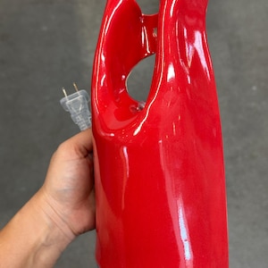 May include: A glossy, bright red ceramic lamp base with a smooth, curved, and abstract sculptural form. The object features a central open cutout and includes a clear electrical cord with a standard two-prong plug extending from the side.