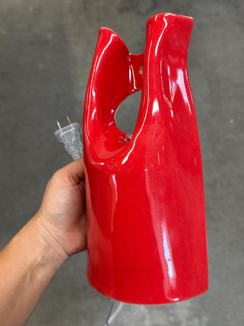 May include: A glossy, bright red ceramic lamp base with a smooth, curved, and abstract sculptural form. The object features a central open cutout and includes a clear electrical cord with a standard two-prong plug extending from the side.