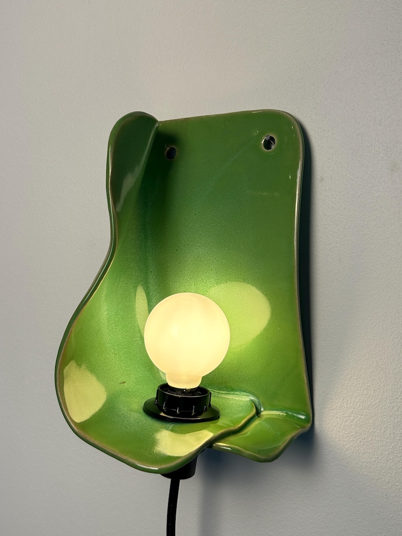 Pu&ograve; includere: Una lampada da parete con una piastra posteriore in ceramica verde lucida dalla forma organica, dotata di due fori di montaggio nella parte superiore. Un portalampada nero sostiene una lampadina rotonda opaca che emette una luce soffusa.