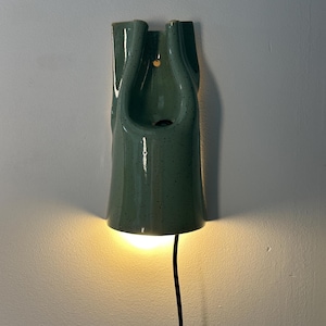 May include: A wall-mounted ceramic light fixture in a speckled sage green glaze. The elongated, vertical form features a central cutout and a small circular hole for mounting. A warm light glows from the bottom opening, with a black power cord extending downward.