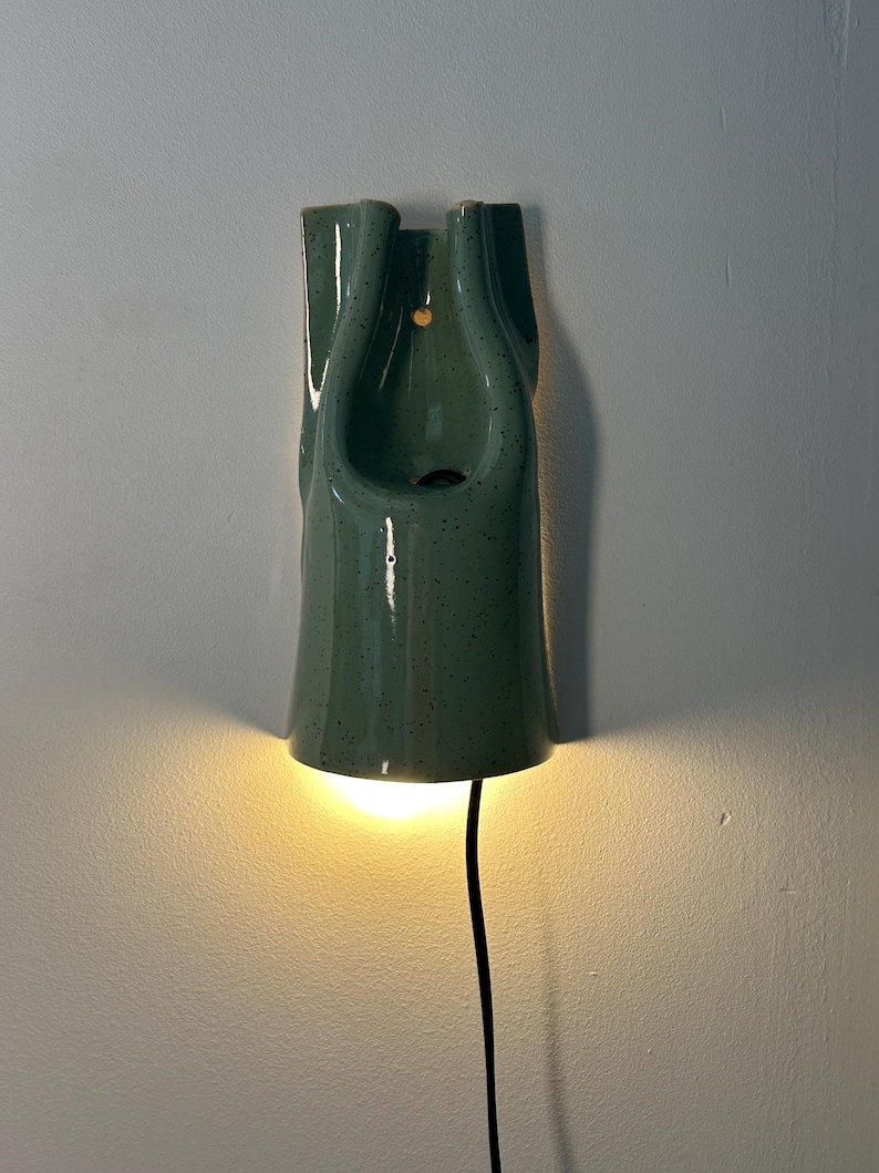 May include: A wall-mounted ceramic light fixture in a speckled sage green glaze. The elongated, vertical form features a central cutout and a small circular hole for mounting. A warm light glows from the bottom opening, with a black power cord extending downward.
