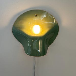 May include: A wall-mounted lamp featuring a glossy, organic, shell-like ceramic shade in a gradient of dark green to light yellow. A single glowing light bulb is centered within the curved form, and a thin, light-colored electrical cord hangs below.