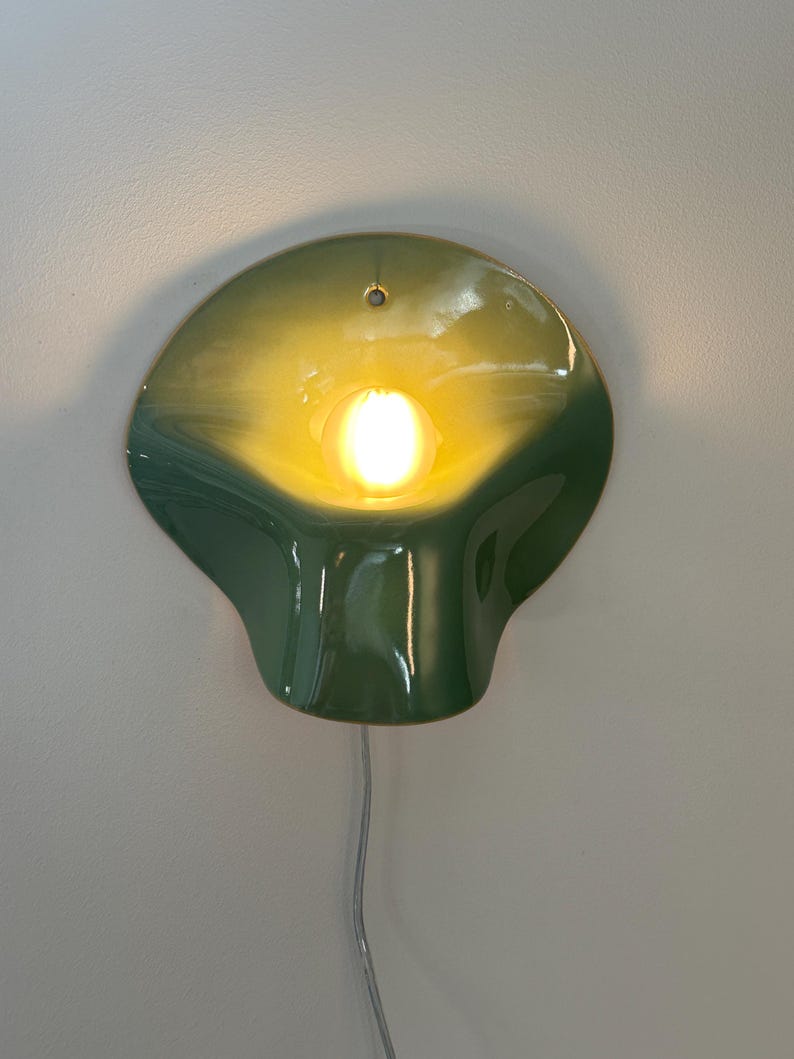 May include: A wall-mounted lamp featuring a glossy, organic, shell-like ceramic shade in a gradient of dark green to light yellow. A single glowing light bulb is centered within the curved form, and a thin, light-colored electrical cord hangs below.