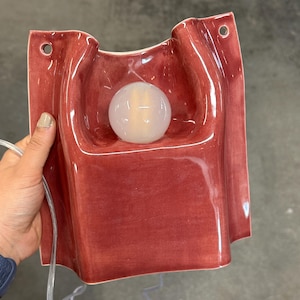 Puede incluir: Una l&aacute;mpara de pared de cer&aacute;mica con un acabado brillante de color rosa intenso y una forma org&aacute;nica ondulada. El aplique cuenta con una bombilla blanca esf&eacute;rica central y dos peque&ntilde;os orificios circulares en las esquinas superiores para su montaje, con un cable transparente.
