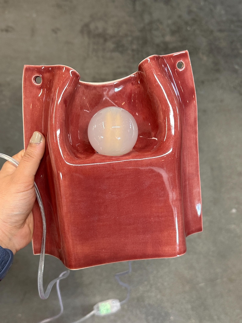 Puede incluir: Una l&aacute;mpara de pared de cer&aacute;mica con un acabado brillante de color rosa intenso y una forma org&aacute;nica ondulada. El aplique cuenta con una bombilla blanca esf&eacute;rica central y dos peque&ntilde;os orificios circulares en las esquinas superiores para su montaje, con un cable transparente.