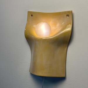 May include: A wall-mounted sconce featuring a curved, yellow ceramic body with a glossy finish. The fixture includes two circular mounting holes at the top corners and a single, spherical, frosted white glass bulb centered in the recessed area.