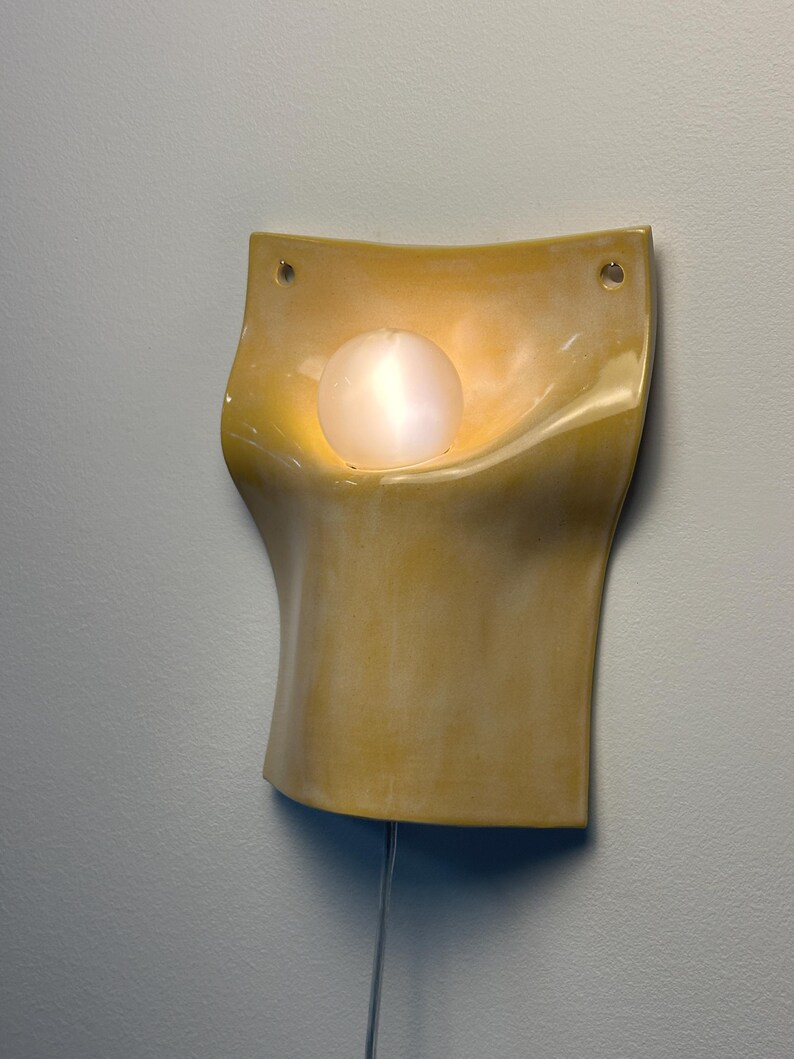 May include: A wall-mounted sconce featuring a curved, yellow ceramic body with a glossy finish. The fixture includes two circular mounting holes at the top corners and a single, spherical, frosted white glass bulb centered in the recessed area.