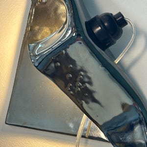 Peut inclure: Une applique murale en c&eacute;ramique avec une finition argent&eacute;e m&eacute;tallique brillante et une forme organique incurv&eacute;e. Le luminaire comprend une douille noire pour ampoule et un cordon &eacute;lectrique transparent sortant de la base, fix&eacute; sur une plaque arri&egrave;re.
