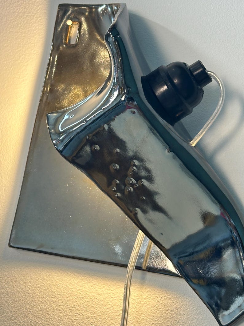 Peut inclure: Une applique murale en c&eacute;ramique avec une finition argent&eacute;e m&eacute;tallique brillante et une forme organique incurv&eacute;e. Le luminaire comprend une douille noire pour ampoule et un cordon &eacute;lectrique transparent sortant de la base, fix&eacute; sur une plaque arri&egrave;re.