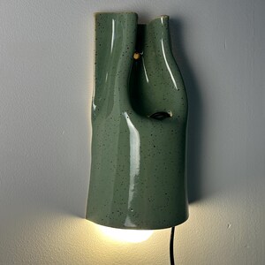 May include: A wall-mounted ceramic light fixture with a sage green speckled glaze. The lamp features an organic, fluid, and asymmetrical shape with a hollow center. A warm light glows from the bottom opening, and a black power cord extends downward.