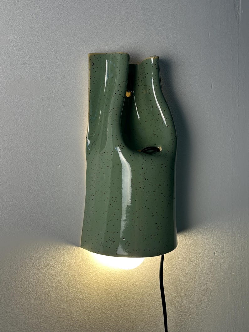 May include: A wall-mounted ceramic light fixture with a sage green speckled glaze. The lamp features an organic, fluid, and asymmetrical shape with a hollow center. A warm light glows from the bottom opening, and a black power cord extends downward.