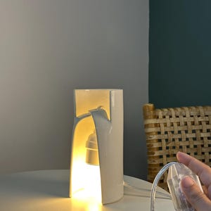 May include: A white ceramic table lamp with a cylindrical, arched design that emits a warm, glowing light from an internal bulb socket. The lamp features a clear power cord with an integrated switch, providing a clean and functional aesthetic.
