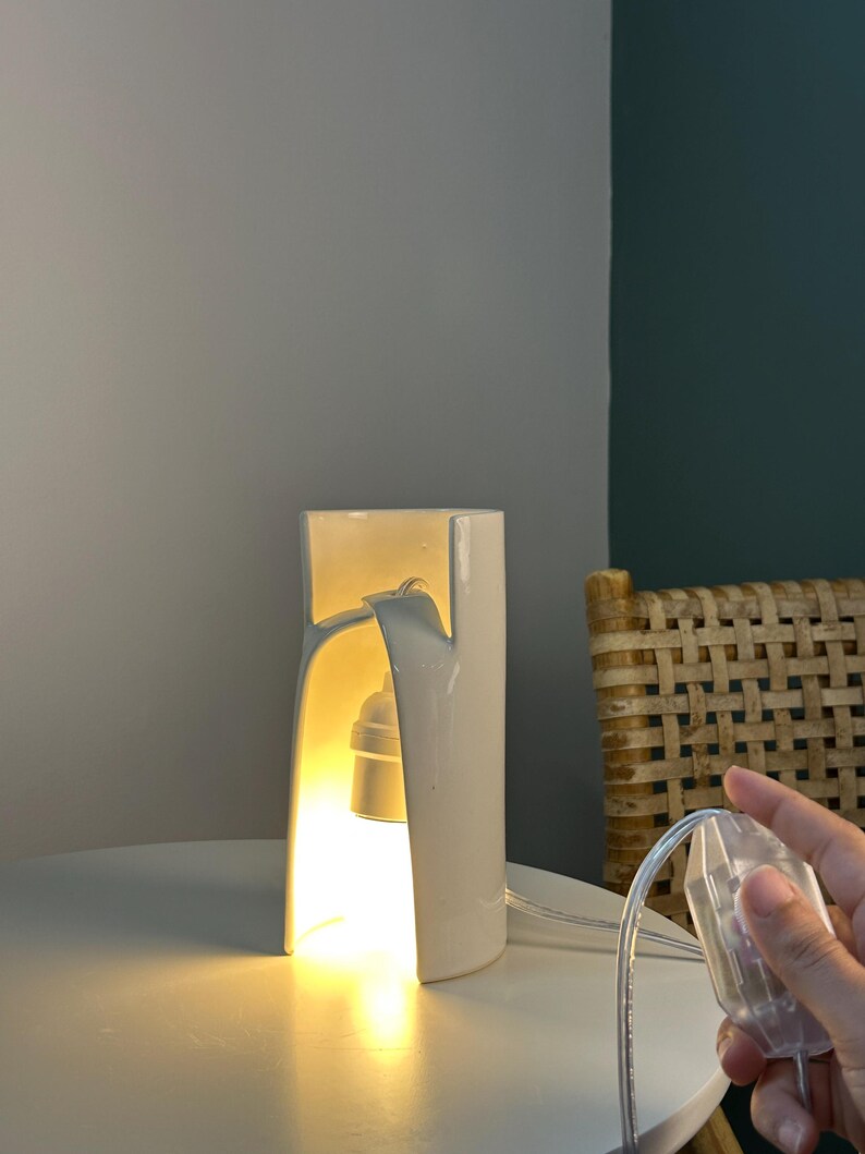 May include: A white ceramic table lamp with a cylindrical, arched design that emits a warm, glowing light from an internal bulb socket. The lamp features a clear power cord with an integrated switch, providing a clean and functional aesthetic.